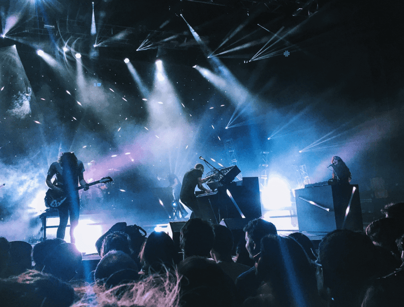 A band performs on stage amidst vibrant lighting effects, with silhouettes of audience members in the foreground.