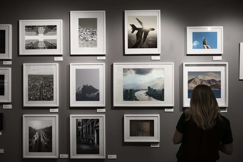 A photograph showing an art exhibition wall featuring multiple framed images in a variety of styles and subjects, including cityscapes, nature, and abstract compositions. A person with long hair is seen from behind, viewing the artwork closely. The room is dimly lit, emphasizing the framed pieces.