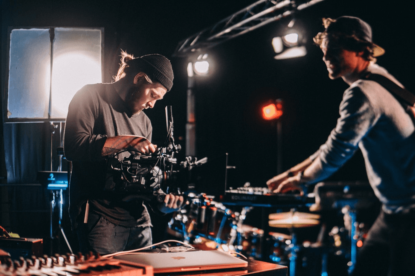 Two individuals are working in a studio setting. The person on the left, wearing a beanie and dark clothing, is focused on adjusting a professional video camera. The person on the right, wearing a backward cap, is interacting with a musical keyboard. The background features studio lights, musical instruments, and a creative workspace environment.